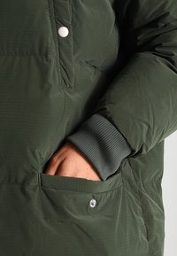 Zielona kurtka puchowa z prążkowanymi mankietami, zapinana na przód na zatrzask oraz z boczną kieszenią. Materiał wydaje się lekki i teksturowany. Zbliżenie na szczegóły.