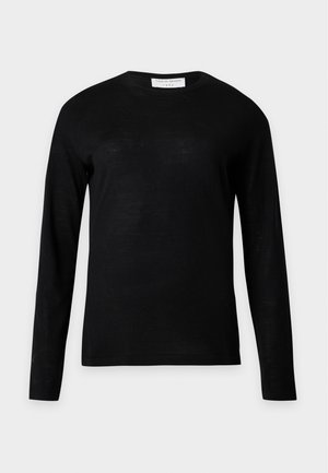 Vrouw met een nauwsluitende zwarte top met lange mouwen, brede beige broek en puntige witte schoenen, staand met één hand in de zak.