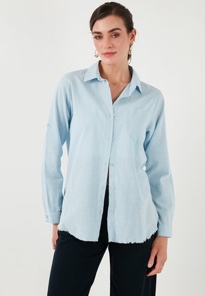 Light blue, long-sleeve shirt in textured fabric with a button-up front, collar, and a single chest pocket. Hem is slightly frayed.