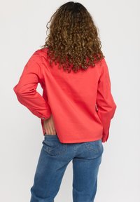 Röd bomullsskjorta med lång ärm, avslappnad passform och knapppdetaljer på ärmsluten. Matchad med blåjeans, sedd bakifrån.