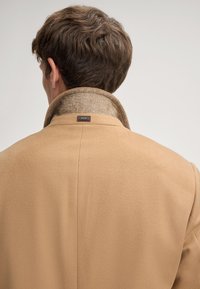 Man met kort bruin haar, gezien van achteren, in een beige jas met een omgeslagen kraag en een JOOP! label op de achterkant.