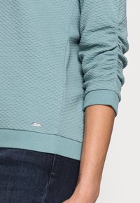 Sweat-shirt bleu clair texturé avec ourlet et poignets côtelés, présentant un motif hexagonal subtil et un petit logo métallique.