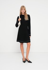 Vestido preto de mangas longas com textura canelada, corpete ajustado, saia evasé e detalhes em branco. Combinado com sapatos pretos de bico fino e uma bolsa branca.