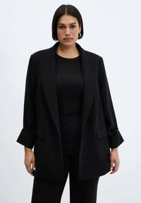 Črna oversize blazer jakna s krojem po meri, žepi in zvitimi rokavi, nošena nad črnim zgornjim delom. Gladek material z formalnim videzom.