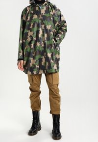 Person klädd i grön, svart och beige kamouflage regnjacka, bruna cargobyxor och svarta snörkängor, står med händerna i fickorna.