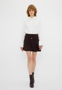 Chemise blanche à manches longues et col classique, associée à une jupe évasée marron avec un accent doré et un détail à la taille.