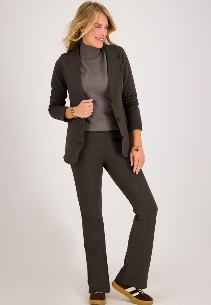 Donkergrijze op maat gemaakte blazer over een lichtgrijze coltrui, gecombineerd met donkergrijze broek en bruine sneakers met witte strepen.