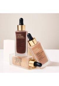 Tre Estee Lauder Futurist SkinTint Serum-flasker i forskjellige nyanser: en mørkebrun og to lysebeige, med gullkapsler og dråpe-topp.