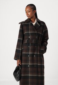 Trench coat de cuadros marrones y negros con botones dobles, cintura ceñida con cinturón y amplia solapa. Modelo sosteniendo un bolso negro.