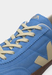 Μπλε αθλητικό παπούτσι από σουέντ με μπεζ κορδόνια, μπεζ λογότυπο "V" στο πλάι και χρυσή σήμανση στη γλώσσα, εμφανιζόμενο από κοντινή γωνία.