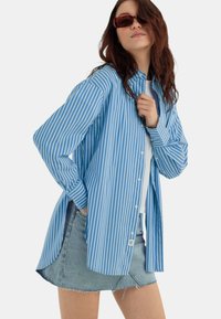 Camicia a righe blu e bianche con bottoni, dal taglio rilassato, abbinata a una mini gonna di jeans chiari con orli sfibrati.