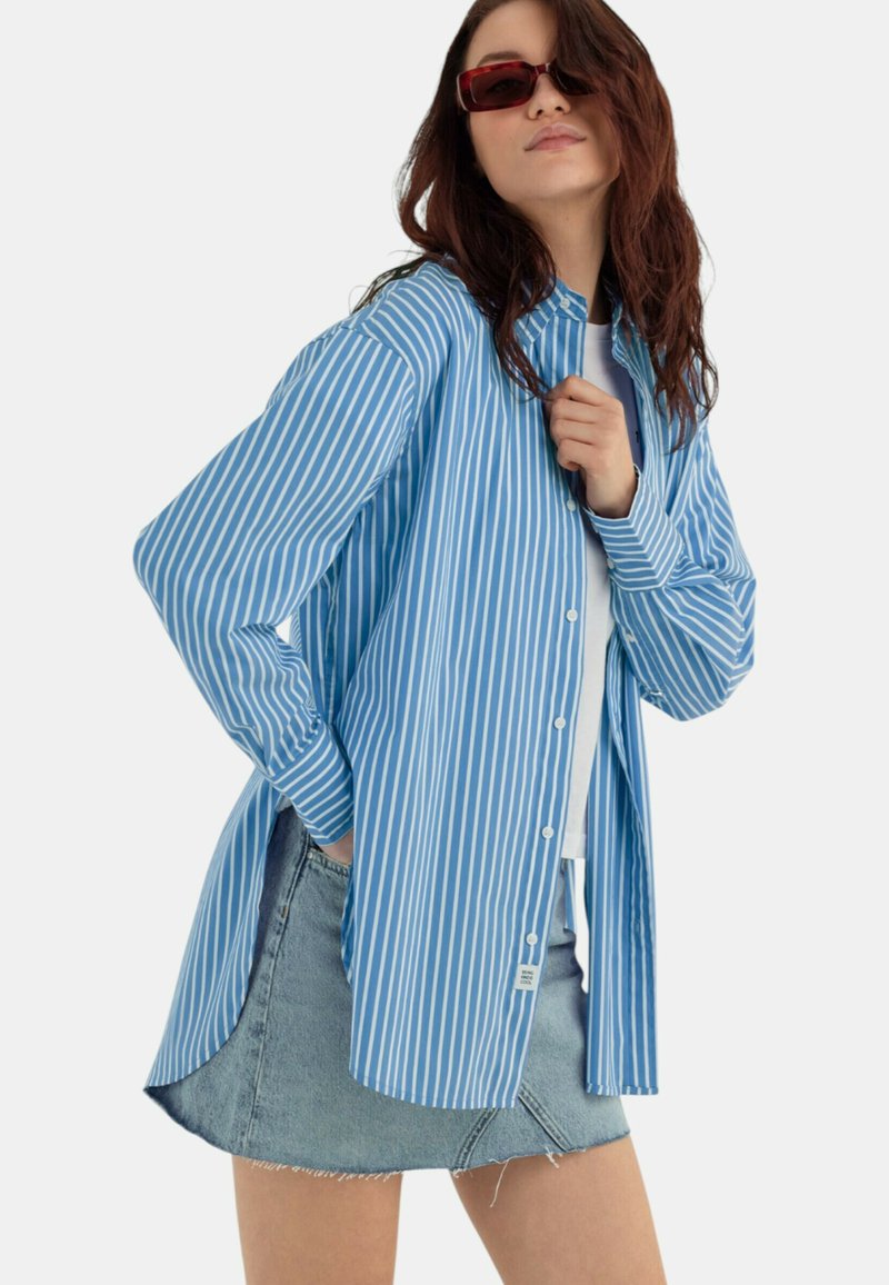 Camicia a righe blu e bianche con bottoni, dal taglio rilassato, abbinata a una mini gonna di jeans chiari con orli sfibrati.