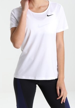 Femme portant un t-shirt blanc Nike à manches courtes et des leggings noirs avec des panneaux bleus, main touchant le cou, debout devant un fond uni.