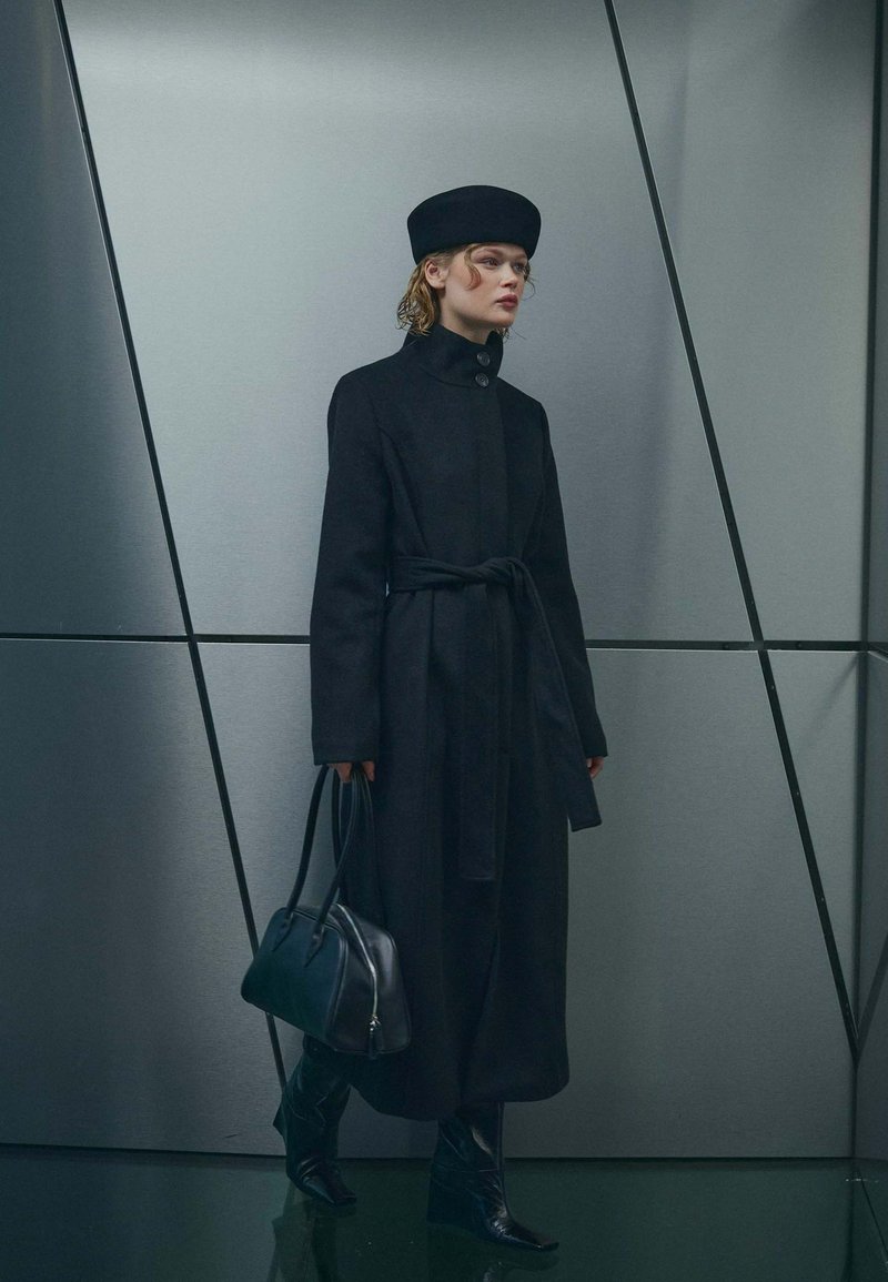 Femme portant un long manteau noir et un chapeau, tenant un sac à main noir, debout contre un mur gris avec des détails de lignes géométriques.