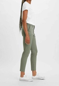 Gröna åtsittande byxor med en slät textur, tillsammans med en vit t-shirt med kort ärm och vita sneakers, sedd från sidan.