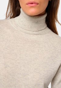 Pull à col roulé beige clair avec une texture côtelée et une coupe ajustée, présentant un col haut et un détail en tissu doux.