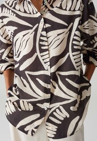 Camicia leggera con bottoni, con un motivo a foglie nero e beige, colletto grande e maniche arrotolate. Presenta tasche frontali e una texture morbida.