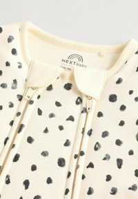 Vêtement pour bébé en coton avec une base crème, présentant des motifs à pois gris, une fermeture éclair et un col arrondi. Étiquette de la marque visible.