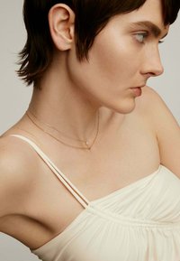 Dos delicados collares superpuestos alrededor del cuello; una cadena de oro presenta un pequeño colgante. El sujeto lleva una blusa sin mangas blanca con tela fruncida.