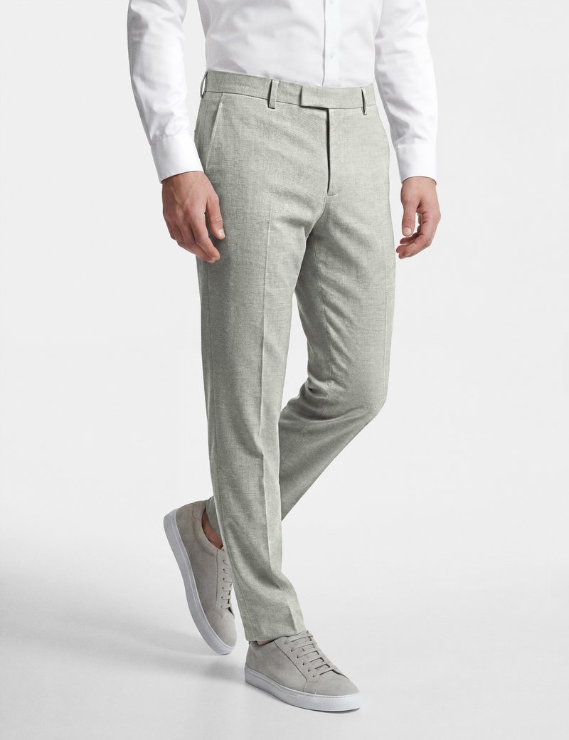 Mann trägt hellgraue Slim-Fit-Hose, weißes Hemd und hellgraue Sneakers, steht mit leicht angewinkeltem Bein.