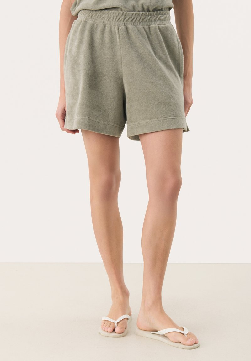 Groene badstof shorts met een elastische tailleband en zijzakken, met een relaxte pasvorm en omgeslagen zoom. Gepaard met witte teenslippers.