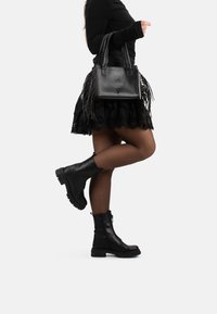 Borsa in pelle nera con frange, caratterizzata da dettagli in rilievo e manico intrecciato, abbinata a una gonna nera in pizzo e stivaletti neri.