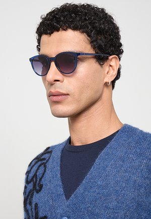 Uomo con capelli corti e ricci che indossa occhiali da sole blu, un cardigan blu a fantasia e un piccolo orecchino a cerchio su uno sfondo chiaro.