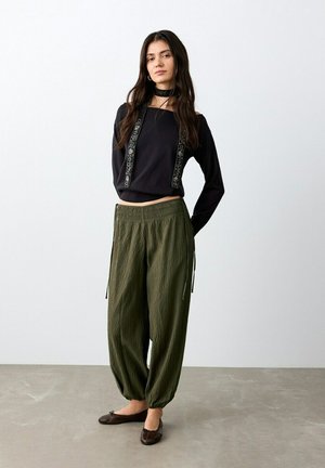 Jeune femme debout portant un haut noir décolleté avec des bretelles brodées, un pantalon ample vert olive et des ballerines marron foncé.