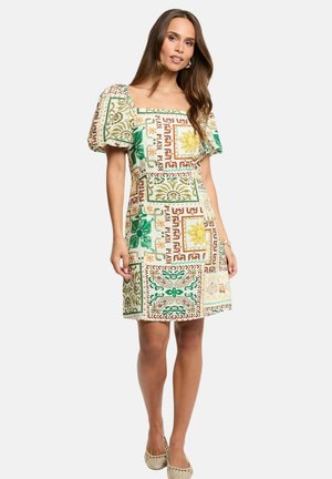 Donna che indossa un vestito fino al ginocchio con scollo quadrato e maniche a sbuffo con motivi geometrici e floreali nei toni del verde, giallo e marrone.