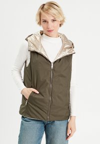 Gilet sans manches vert olive avec col doré brillant, fermeture zippée et poches latérales. Porté par-dessus un haut à manches longues blanc, coupe décontractée.
