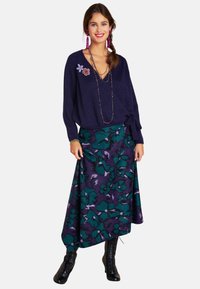 Navyblauer V-Ausschnitt-Pullover mit einer Bindeschleife, kombiniert mit einem ausgestellten Rock mit einem grünen und lila Blumenmuster. Tragen Sie dazu schwarze Schnürstiefel.