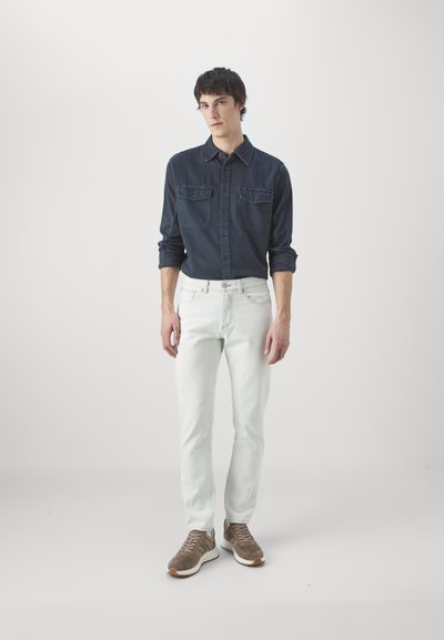 Mørkeblå denimskjorte med to brystlommer, kombinert med lyse jeans og brune sneakers, satt mot en nøytral bakgrunn.