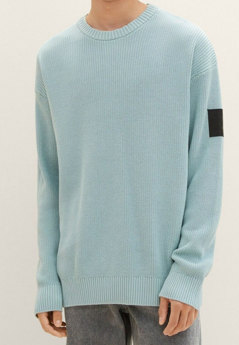 Osoba v světle modrém pleteném svetru s žebrovanými manžetami a spodním lemem, kulatým výstřihem a černou nášivkou na levém rukávu.