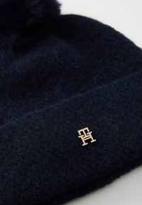 Navyblauwe wollen gebreide beanie met een nep-bont pompon en een gouden logo patch aan de voorkant. Zachte textuur met ribbel details.