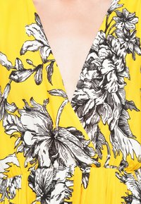 Gul stof med sort blomstermønster, der har en V-udskæring og en samlet talje for en svævende silhuet.