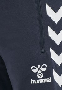 Pantalones jogger de color navy con un bolsillo con cremallera, un patrón de chevron blanco en contraste y un logo en el muslo en blanco. Tela suave y elástica.