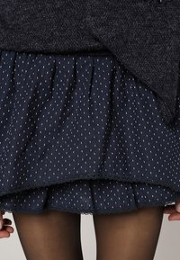 Námořnická modrá sukně s texturovaným vzorem bílých vertikálních pruhů, s volánkovým lemem a krajkovým zdobením. V kombinaci s tmavými punčochami.