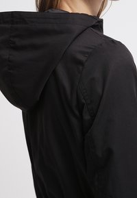 Black hooded jacket with a smooth, matte texture. Features a detailed seam at the shoulder and a relaxed fit, emphasizing functionality.