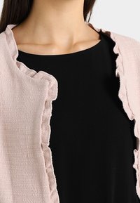 Cardigan abierto de color rosa claro con una tela texturizada y bordes con volantes, llevado sobre una blusa negra lisa.