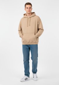 Ung man som står med händerna i fickan, iklädd en beige huvtröja, blå jeans och vita sneakers med svarta ränder, mot en vit bakgrund.