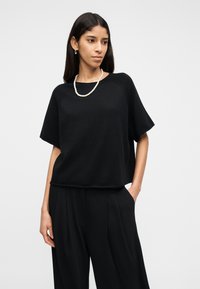 Femme portant un haut noir à manches courtes ample et un pantalon noir à jambes larges, accessoirisée d'un collier de perles et de boucles d'oreilles argentées en forme de créoles.