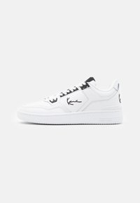 White leather sneaker with black accents, featuring a signature logo on the side, round toe, and textured rubber sole.