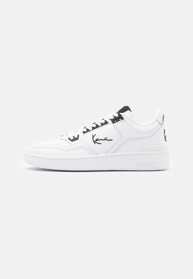 White leather sneaker with black accents, featuring a signature logo on the side, round toe, and textured rubber sole.