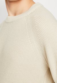 Ljusbeige stickad tröja med ett strukturerat mönster, ribbad rundhals och raglanärmar. Mjuk material syns i detalj.