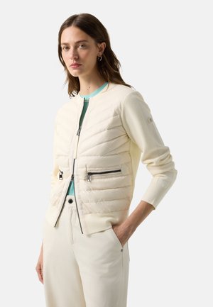 Kvinde iført cremet quiltet jakke med lynlåslommer over lyseblå top, cremefarvede bukser, stående med den ene hånd i lommen.