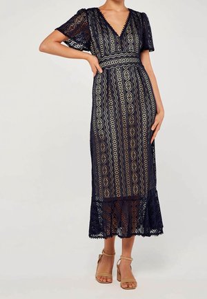 Femme portant une robe midi en dentelle bleu marine avec manches courtes, col en V, taille cintrée, et des sandales à talons hauts avec bracelet à la cheville couleur beige.