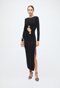 Zwarte jurk met lange mouwen, voorzien van uitsparingen met ronde accenten, een zijsplit en een nauwsluitend silhouet. Model houdt een gouden metallic clutch vast.