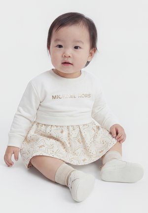 Bambino piccolo con capelli scuri seduto, indossando un maglione bianco con il testo "Michael Kors", gonna a fantasia, calzini chiari e scarpe bianche su sfondo bianco.