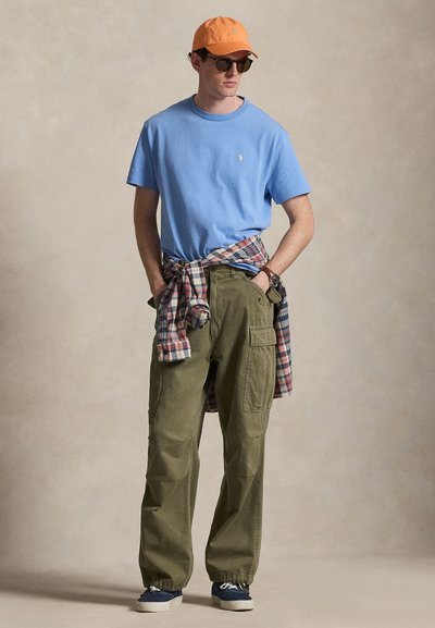 Άνδρας που φοράει πορτοκαλί καπέλο, γυαλιά ηλίου, γαλάζιο μπλουζάκι, καρό πουκάμισο δεμένο στη μέση, χακί παντελόνι τύπου cargo και ναυτικά αθλητικά παπούτσια, στέκεται με τα χέρια στις τσέπες.