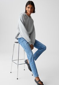 Donna seduta su uno sgabello di metallo, indossa un hoodie grigio, jeans azzurri chiari e scarpe slip-on nere con fibbia.